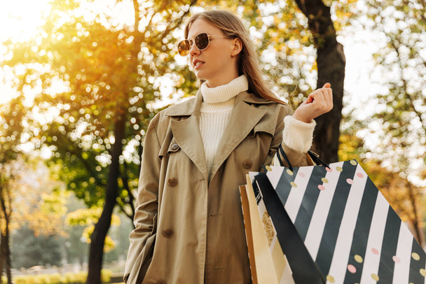 stylish woman in a beige raincoat and sun goggles with shopping bags in her hands, on against the autumn trees, season of discounts, sale. business woman shopping in city - 写真・画像