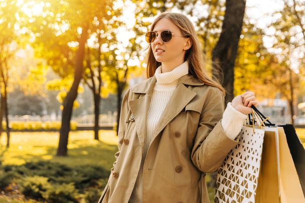 stylish woman in a beige raincoat and sun goggles with shopping bags in her hands, on against the autumn trees, season of discounts, sale. business woman shopping in city - Foto, imagen