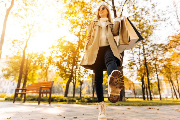 close-up of a woman's autumnal shoes, walking, a woman holding shopping bags on the street, in the park, trees are falling, yellow leaves are falling. woman in beige raincoat and autumn boots - Foto, afbeelding