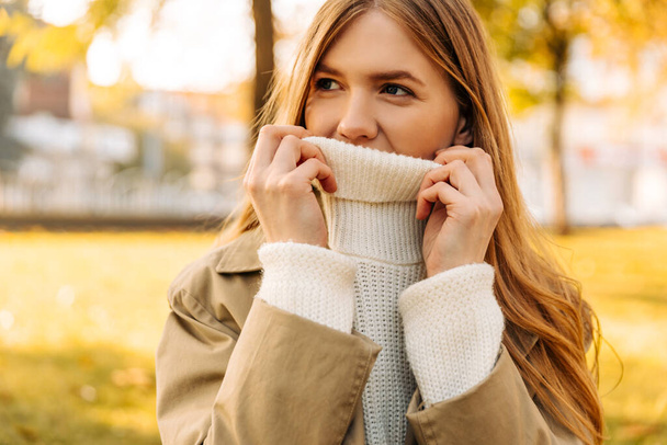 beautiful blonde, holds the neck of the sweater, pulls it up on her face. a woman holds her sweater with her hands, in a beige raincoat, against the background of an autumn park - Fotoğraf, Görsel