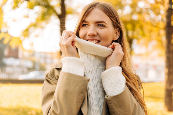 beautiful blonde, holds the neck of the sweater, pulls it up on her face. a woman holds her sweater with her hands, in a beige raincoat, against the background of an autumn park - Foto, imagen