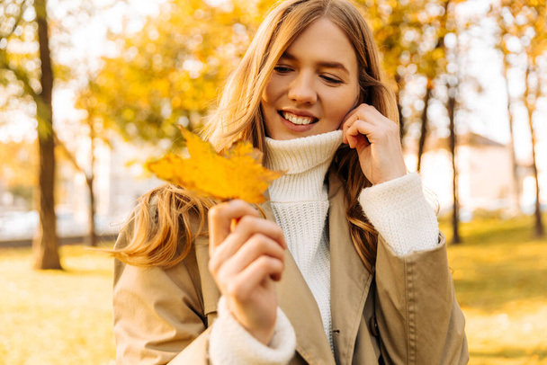 beautiful woman in a beige raincoat shows a yellow leaf that has fallen from a tree. autumn weather, mood. blonde dreams in the park, autumn weather - Photo, image