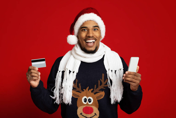 スマートフォンとクレジットカードのある陽気な黒人男性はクリスマスを祝う - 写真・画像