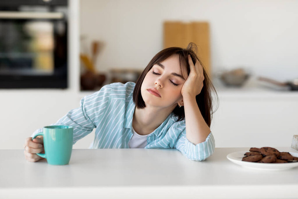 Νυσταγμένη νεαρή γυναίκα που πίνει καφέ, αισθάνεται κουρασμένη, υποφέρει από αϋπνία και διαταραχή ύπνου, κάθεται στην κουζίνα - Φωτογραφία, εικόνα