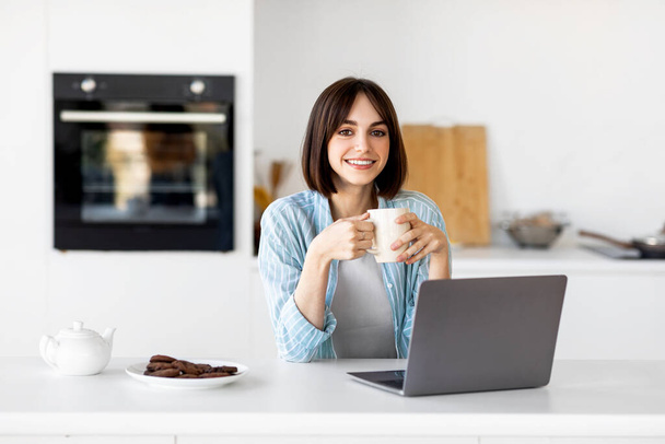 Mutlu bir kadının portresi dizüstü bilgisayar ve bardak kullanması, sabahları sıcak kahve içmesi, modern mutfakta oturması. - Fotoğraf, Görsel