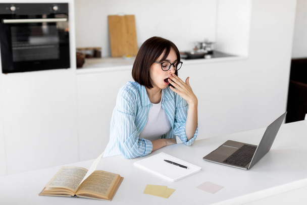 Tired sleepy lady yawning while working or studying on laptop online from home, sitting in modern kitchen, free space - 写真・画像