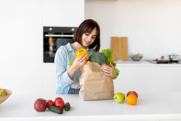 Tisíciletá žena vybaluje papírový sáček s čerstvou organickou zeleninou a ovocem, stojící v moderní kuchyni - Fotografie, Obrázek