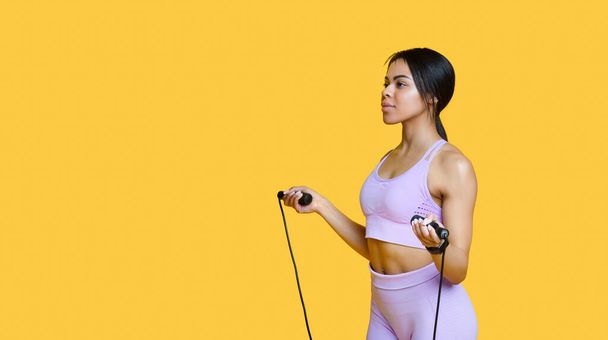 ジャンプロープを持つスポーティアフリカ系アメリカ人女性,スポーツウェアを着て、フィットネストレーニングの準備ができて,黄色の背景 - 写真・画像