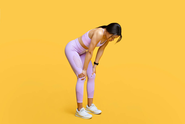Deporte, respira y descansa. Mujer negra cansada tomando descanso en el entrenamiento, apoyada en las rodillas, fondo amarillo - Foto, imagen
