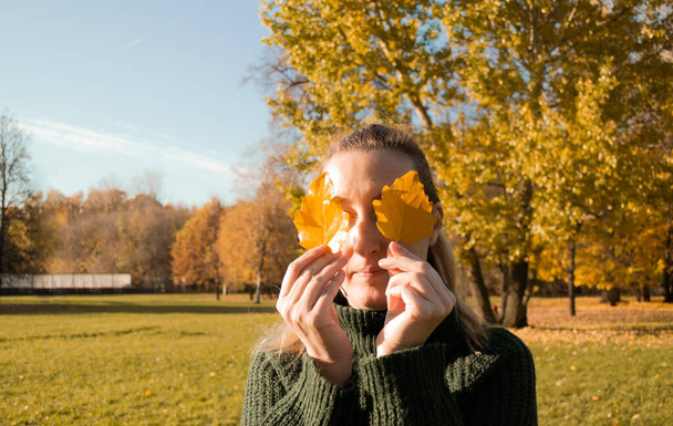 Gözleri kapalı kadın portresi iki sarı yaprak tutar, elleri yakın, arka plan sonbahar ağaçları. - Fotoğraf, Görsel