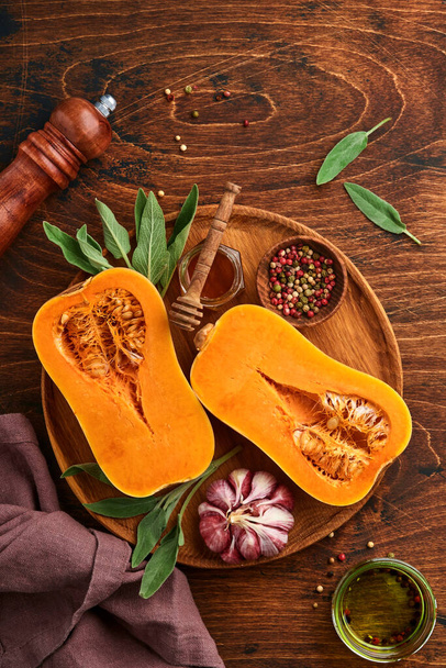 Halves of raw organic butternut squash with sage leaf, multicolored pepper garlic, honey, salt and pepper on old wooden background. Food background. Top view with copy space. - Foto, Bild