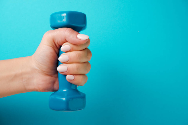 Fitness concept A female hand holds a blue plastic dumbbell. Blue background. - Photo, Image