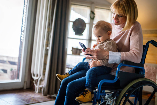 Menschen Technologie Familienglück Konzept. Glückliche Mutter mit Behinderung im Rollstuhl mit ihrem Kind mit digitalem Tablet zu Hause - Foto, Bild