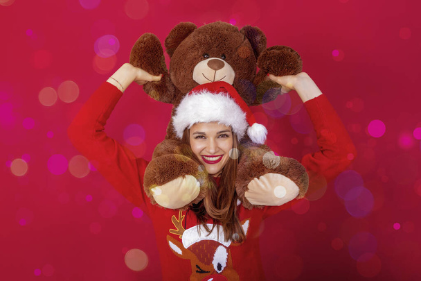Lächelnde hübsche Frau mit Weihnachtsmann-Hut, Rentier-Pullover und riesigem Teddybär auf den Schultern. - Foto, Bild