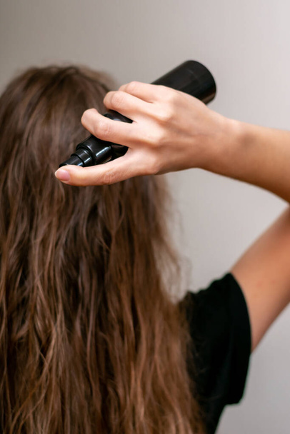 Young woman hand holds bottle and spraying her wavy blond long hair for making it silken and strong. Treatment for hair loss and care. - Foto, imagen