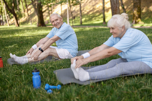 Positive senior man does head to knee exercise with wife sitting on meadow - Photo, Image