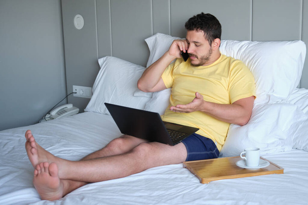Hombre hablando por teléfono y trabajando con el ordenador portátil en una cama de hotel. - Foto, imagen
