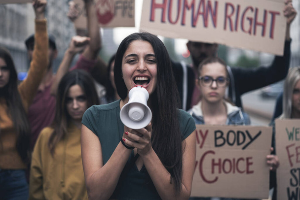 Group of young people protesting on the street with signs raised to promote human rights. Focus on the woman in front screaming to the megaphone - Photo, Image