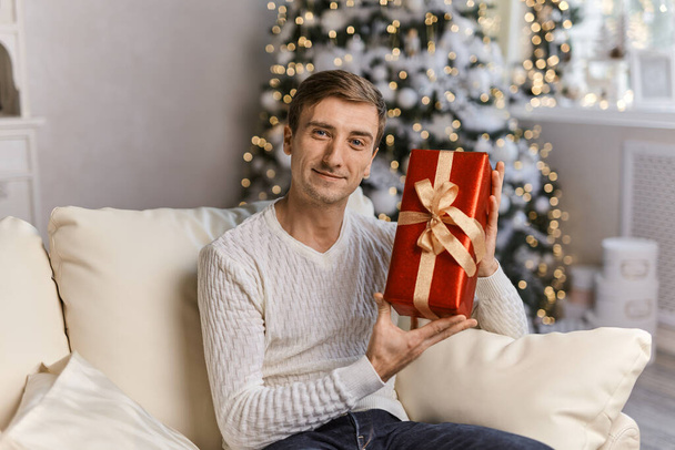 Счастливый молодой человек держит подарок в рождественское время. улыбающийся мужчина в зимнем свитере с красной подарочной коробкой
 - Фото, изображение