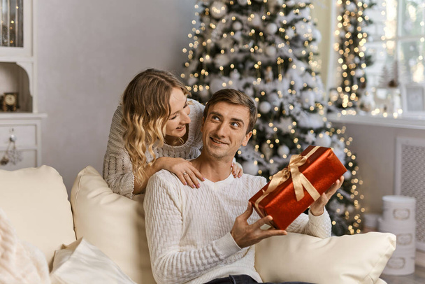 Gyönyörű pár, akik együtt várják az új évet, miközben ajándékdobozzal a kezükben ölelkeznek, közel a gyönyörű karácsonyfához, a hangulatos nappali kanapéján, megvilágított koszorúval. - Fotó, kép