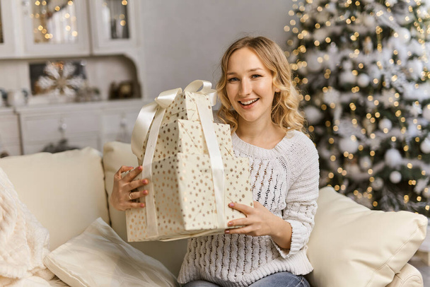 Weihnachts- und Neujahrsgeschenke. Glückliche junge Dame hält und umarmt Geschenkbox, sitzt auf dem Sofa zu Hause im Wohnzimmer - Foto, Bild