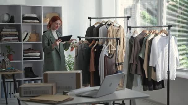 tienda minorista, una joven empresaria sosteniendo una tableta en la mano toma un inventario de la ropa en los estantes en una boutique - Imágenes, Vídeo