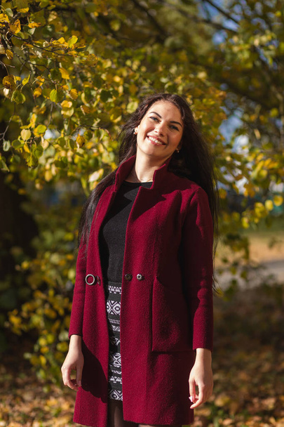 Portrait of a joyful young woman enjoying in the autumn park. Beautiful brunette girl in autumn red coat and black sweater. Relax in nature. High quality photo - Foto, immagini
