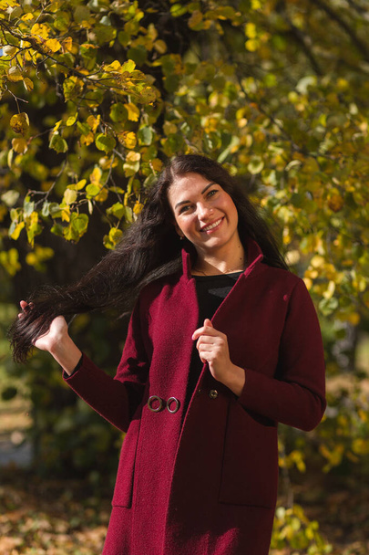 Portrait of a joyful young woman enjoying in the autumn park. Beautiful brunette girl in autumn red coat and black sweater. Relax in nature. High quality photo - Φωτογραφία, εικόνα