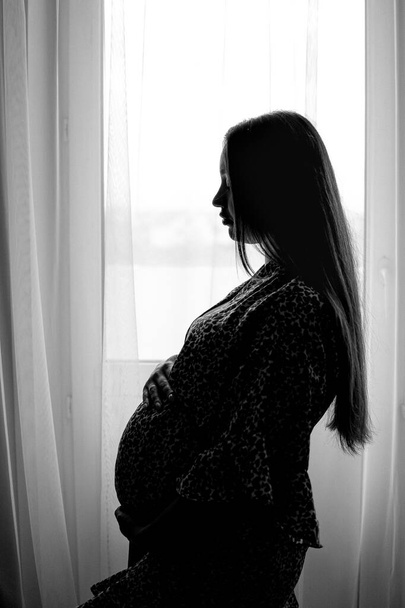 Pregnant woman silhouette photo. Pregnant female standing near the window. Side-view portrait of pregnant woman. Selective focus. Film grain. - Photo, image