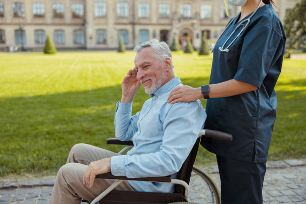 Happy aged man, recovering patient in a wheelchair on a walk with a nurse wearing face shield and mask on a summer day outdoors - Foto, afbeelding