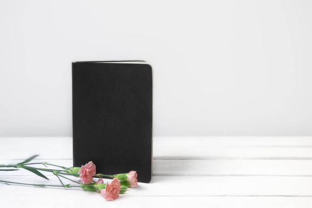 Białe drewniane biurko z czarną pustą książką lub notatnikiem i różowe kwiaty. Z przodu. Miejsce na tekst - Zdjęcie, obraz