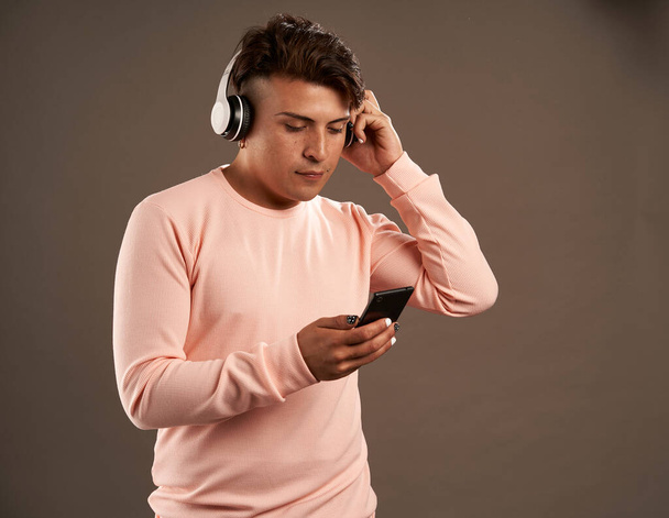 latynoski mężczyzna ze słuchawkami patrzący na telefon komórkowy słuchający muzyki odizolowanej na szarym tle z różową odzieżą kopiuj przestrzeń - Zdjęcie, obraz