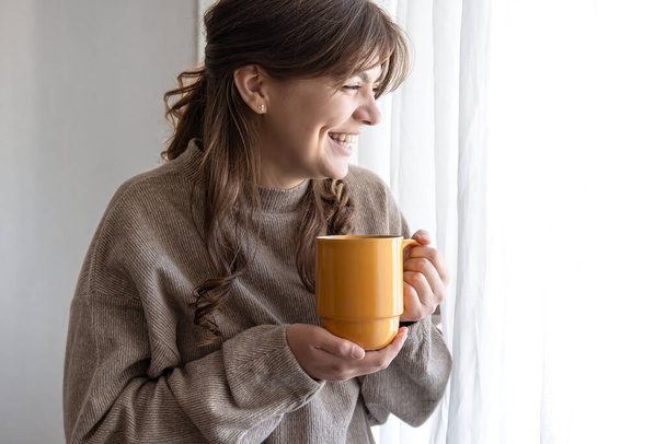 Привлекательная молодая женщина с чашкой горячего напитка у окна. - Фото, изображение