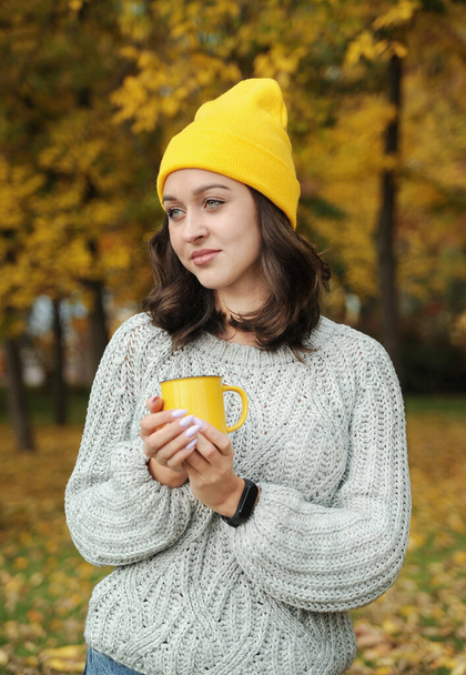 Sonbahar parkında elinde fincanı tutan sarı şapkalı güzel bir kadın. Genç bayan dışarıda sıcak kahve içiyor. Sonbahar mevsimi konsepti. - Fotoğraf, Görsel