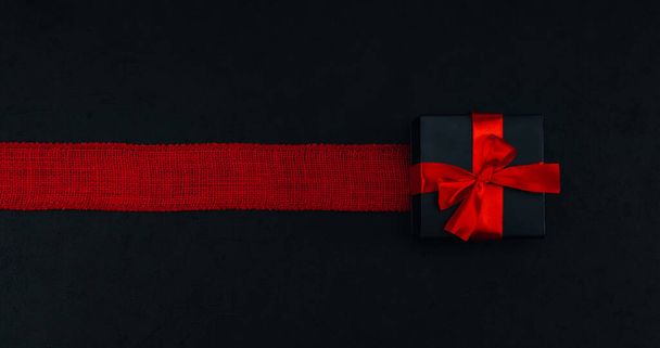Červená dárková krabice s černou stuhou a pytlovitým pruhem leží uprostřed na černém pozadí s prostorem pro text, zblízka. Valentýnský koncept, černý pátek a Vánoce. - Fotografie, Obrázek