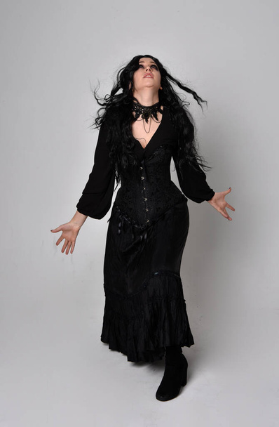 Plně dlouhý portrét tmavovlasé ženy v černém viktoriánském kostýmu čarodějnice s korzetem, stojící póza gesturálními pohyby rukou, na pozadí ateliéru. - Fotografie, Obrázek