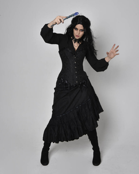 Retrato de comprimento total de mulher de cabelos escuros vestindo traje de bruxa vitoriana preta com espartilho, postura em pé com movimentos gestuais das mãos, contra o fundo do estúdio. - Foto, Imagem