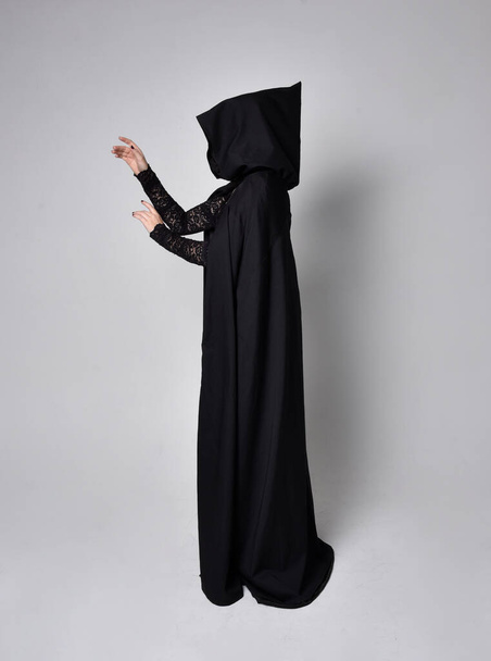 Портрет темноволосої жінки в чорному костюмі вікторіанської відьми з плащем і гострим капелюхом. стояча поза, вид ззаду, з рухами рук жестів, на фоні студії
. - Фото, зображення