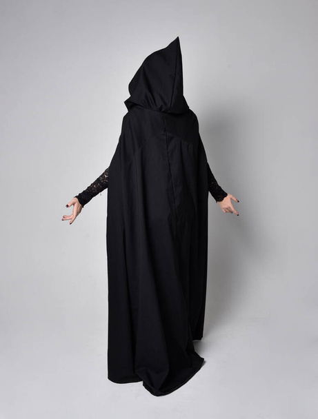 Portrait complet de femme aux cheveux foncés portant un costume de sorcière victorienne noire avec un manteau et un chapeau pointu. pose debout, vue de dos, avec mouvements gestuels des mains, sur fond de studio. - Photo, image