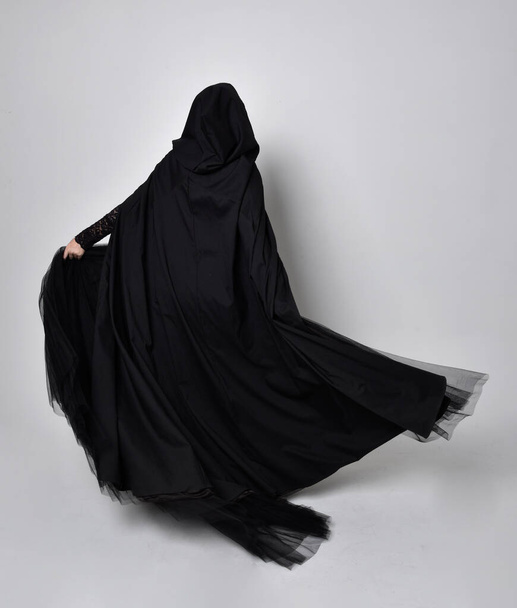 Полноразмерный портрет темноволосой женщины в чёрном викторианском костюме ведьмы в плаще и остроконечной шляпе. Стоя поза, вид сзади, с жестом движения рукой, на фоне студии. - Фото, изображение