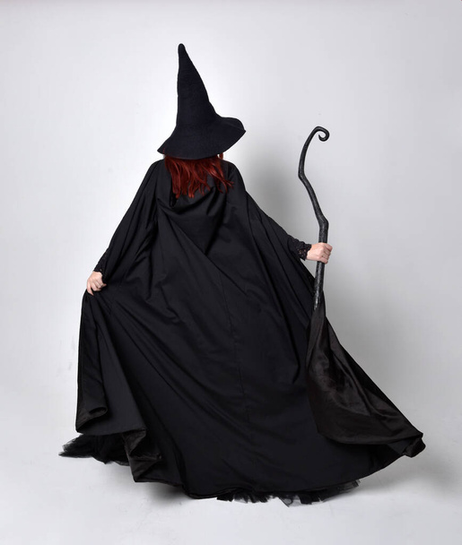Полноразмерный портрет темноволосой женщины в чёрном викторианском костюме ведьмы в плаще и остроконечной шляпе. Стоя поза, вид сзади, с жестом движения рукой, на фоне студии. - Фото, изображение