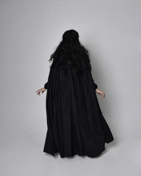 Portrait complet de femme aux cheveux foncés portant un costume de sorcière victorienne noire avec un manteau et un chapeau pointu. pose debout, vue de dos, avec mouvements gestuels des mains, sur fond de studio. - Photo, image