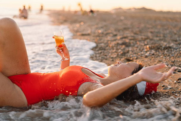 Young beautiful woman in a Santa hat with a glass of champagne in her hands walk on the beach. - Foto, Imagen