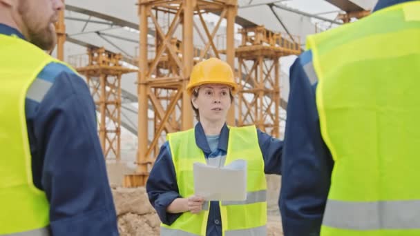 Ruční záběr ženského nadřízeného v bezpečnostní vestě a pevném klobouku s dokladem a bezpečnostní pokyny mužským pracovníkům na staveništi - Záběry, video