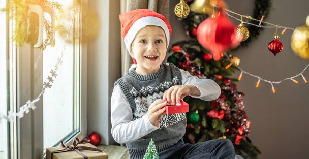 Cute little boy in a red Santa hat is sitting by the window in a decorated room and holding a small open gift box in his hands and smiling. Concept of a festive New Year and Christmas mood. - Фото, изображение