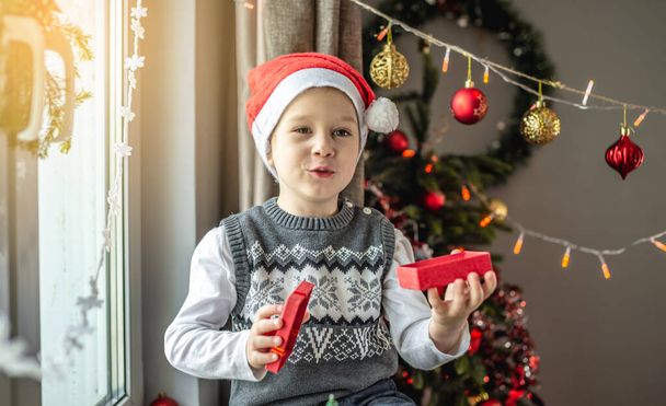Cute little boy in a red Santa hat is sitting by the window in a decorated room and holding a small open gift box in his hands . Concept of a festive New Year and Christmas mood. - Photo, image