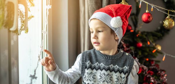 Cute little boy in a red Santa hat is sitting at the window next to the Christmas tree and looking at the festive decorations at home. Concept of New Year atmosphere and holiday mood. - Photo, Image