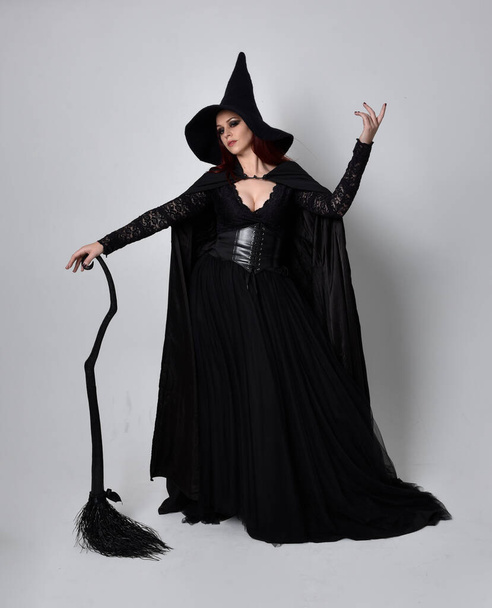 Full length portrait of dark haired woman wearing  black victorian witch costume.  standing pose, holding a broomstick  with  gestural hand movements,  against studio background. - Φωτογραφία, εικόνα