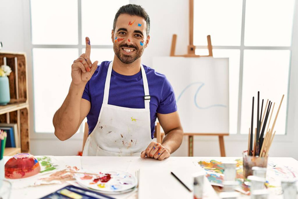 Jovem hispânico com barba no estúdio de arte com rosto pintado mostrando e apontando para cima com o dedo número um enquanto sorri confiante e feliz.  - Foto, Imagem