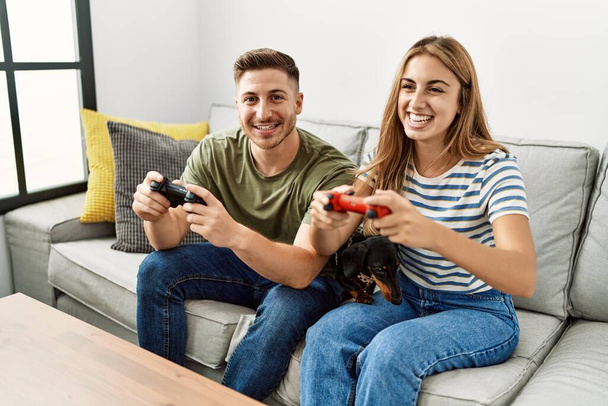 Junges hispanisches Paar lächelt glücklich beim Videospiel auf dem Sofa sitzend mit Hund zu Hause. - Foto, Bild